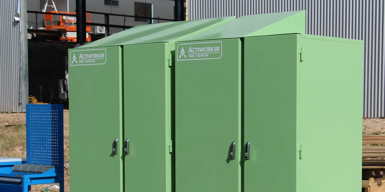 The image shows two large, green, industrial storage lockers placed on wooden pallets outdoors. The lockers have secure doors with handles and are labeled with "ACTIWORK." There is also a blue mobile tool cart with drawers and shelves next to the lockers.